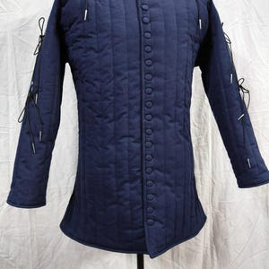 May include: A dark blue quilted doublet featuring vertical stitching, a central row of fabric-covered buttons, and long sleeves with black lace-up ties at the shoulders and elbows. The garment has a high collar and a structured, fitted design.
