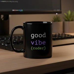 Op de afbeelding: Zwarte keramische mok met de woorden "good vibe {coder}" in wit, paars en groen. De mok staat op een houten bureau met een toetsenbord en computermonitor op de achtergrond. De monitor toont code.