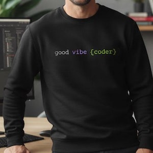 May include: Black sweatshirt with the text "good vibe {coder}" in a gradient of green and purple. The long-sleeved sweatshirt is worn with dark blue trousers. The background includes a computer monitor, desk, and a potted plant.