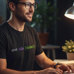 Peut inclure: Un t-shirt noir avec le texte "good vibe {coder}" dans un dégradé de couleurs. La personne porte des lunettes et est assise à un bureau avec un clavier et une souris d'ordinateur. Une lampe de bureau et de petites plantes en pot sont également visibles.