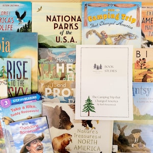 May include: A collection of colorful children's books with titles like "Explore! America's Wildlife," "National Parks of the U.S.A.," and "The Camping Trip That Changed America." A white booklet with a green tree graphic is in the center.