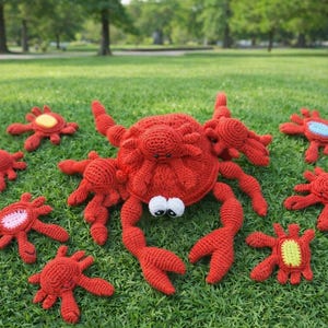 Puede incluir: Una colección de cangrejos rojos de ganchillo hechos a mano de varios tamaños, dispuestos sobre un césped verde. El cangrejo más grande tiene ojos blancos. Los cangrejos más pequeños tienen acentos coloridos. La escena está ambientada al aire libre con árboles al fondo.