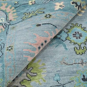 May include: A folded section of a blue rug with a detailed floral pattern. The rug features a light blue background with pink, green, and darker blue floral and geometric designs. The rug's texture is visible.