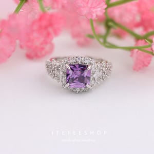 May include: A silver ring with a square, purple gemstone at the center, surrounded by smaller, clear stones. The ring has a detailed, textured band. Pink flowers are in the background. The text "ITEFEESHOP" and "Handcrafted Jewellery" are at the bottom.