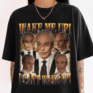Puede incluir: Camiseta negra con un diseño gráfico dorado. El diseño presenta el texto "WAKE ME UP! I CAN'T WAKE UP!" y múltiples imágenes de un hombre con traje y auriculares.