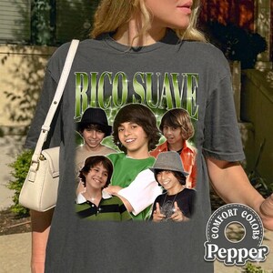 May include: Dark gray t-shirt featuring a graphic of a group of young men with the text "Rico Suave" in green. The shirt has a vintage style and the Comfort Colors Pepper logo is visible on the bottom right.