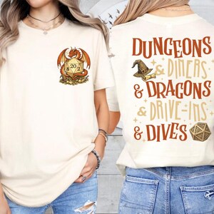 May include: Cream-colored t-shirt featuring a dragon and a 20-sided die graphic. The back of the shirt displays the text "DUNGEONS & DINERS & DRAGONS & DRIVE-INS & DIVES" with a wizard hat and die graphic.