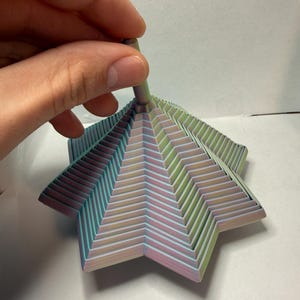 May include: A multi-layered, star-shaped paper sculpture in pastel colors of pink, green, and blue. The sculpture is held by a hand, showcasing its intricate, zig-zag design. The paper art piece has a central, cylindrical stem.