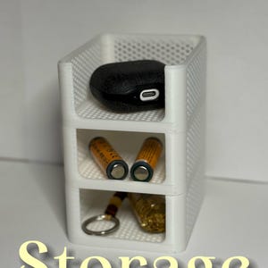 May include: White, three-tiered storage organizer with a mesh design. The top shelf holds a black, oval-shaped item. The middle shelf contains two orange and silver batteries. The bottom shelf has a keychain and a gold-colored item. The word "Storage" is at the bottom.