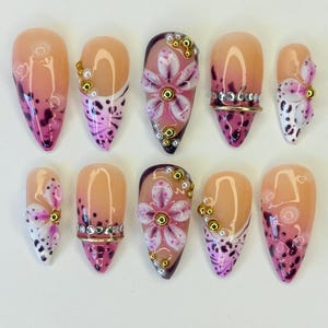 Puede incluir: Un conjunto de diez uñas artificiales con una variedad de diseños. Algunas uñas presentan un diseño floral rosa y morado, mientras que otras tienen un patrón de manchas rosas y moradas. Varias uñas están adornadas con adornos dorados y plateados.