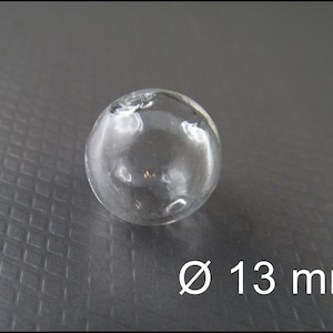 May include: A clear glass sphere with a diameter of 13 mm. The sphere is sitting on a gray textured surface.