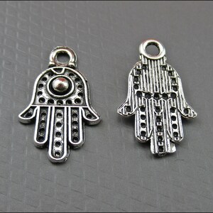 May include: Two silver Hamsa hand charms with intricate designs. The charms have a hole at the top for hanging.