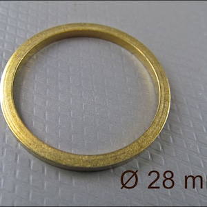 Puede incluir: Un anillo circular de color latón, con un diámetro de 28 mm. El anillo tiene un perfil plano y una superficie ligeramente texturizada. La imagen es un primer plano, destacando el brillo metálico del anillo.