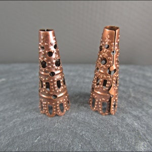 May include: Two copper cone-shaped bead caps with a decorative, openwork design. The caps have a textured surface and a scalloped edge.