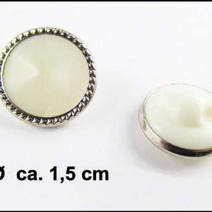 Puede incluir: Dos botones blancos con un borde de metal plateado y un diseño de cuerda. Los botones tienen un diámetro de aproximadamente 1,5 cm.