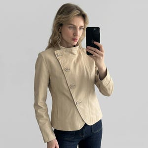May include: A tan leather jacket with a unique asymmetrical button closure and a high, rounded collar. The jacket has long sleeves and a tailored fit. The model is wearing the jacket with dark blue jeans.