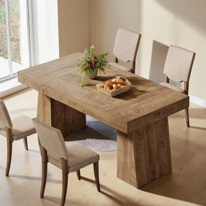 May include: A rectangular wooden dining table with a thick top and two solid pedestal bases, accompanied by four chairs featuring light beige fabric upholstery on the seats and backrests, with a small basket of bread and a green plant on top.