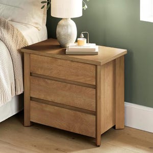May include: A light brown wooden nightstand featuring three horizontal drawers with recessed handles. The top surface holds a ceramic table lamp with a white drum shade and a stack of two rectangular books with a small candle on top.