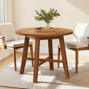 May include: A round wooden dining table with four angled legs and a cross-brace support, accompanied by two armchairs featuring light beige upholstered seats and backrests. A round woven tray sits on the table holding a white vase with greenery.