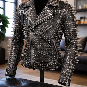 May include: Black leather jacket adorned with silver spikes and studs. The jacket has a classic biker design, featuring a notched collar, an asymmetrical zip, and several pockets. The sleeves have zip cuffs and shoulder straps with buckles.
