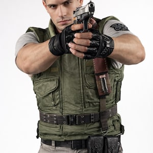Könnte beinhalten: Eine Person in einer grünen taktischen Weste, einem grauen Hemd und Handschuhen, die eine silberne Handfeuerwaffe hält. Die Weste hat mehrere Taschen und einen Gürtel mit Taschen. Die Person schaut direkt in die Kamera.