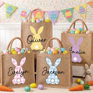 May include: Four burlap Easter tote bags with bunny designs in yellow, pink, blue, and purple. Each bag has a name printed on it: Oliver, Evelyn, Jackson, and Amelia. The bags are filled with colorful Easter eggs.