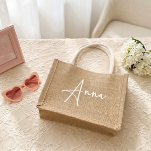 May include: A small, rectangular burlap tote bag with white rope handles. The name "Anna" is written in cursive across the front. Pink heart-shaped sunglasses and a pink picture frame are next to the bag.
