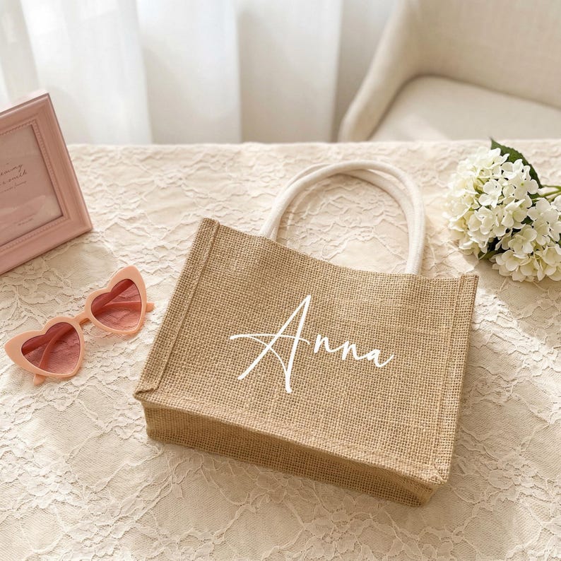 May include: A small, rectangular burlap tote bag with white rope handles. The name "Anna" is written in cursive across the front. Pink heart-shaped sunglasses and a pink picture frame are next to the bag.