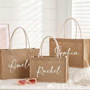 May include: Three burlap tote bags with white handles and personalized names in cursive: Amelia, Rachel, and Sophia. The bags are on a white surface with a pink framed picture and heart-shaped sunglasses.