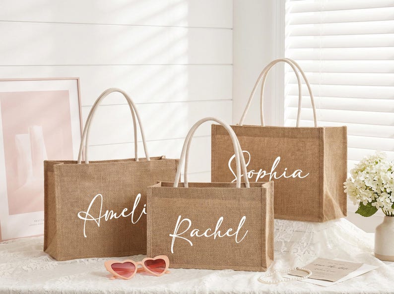 May include: Three burlap tote bags with white handles and personalized names in cursive: Amelia, Rachel, and Sophia. The bags are on a white surface with a pink framed picture and heart-shaped sunglasses.