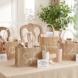 May include: Several burlap tote bags with white handles are arranged on a table. Each bag is personalized with a name in white script. The bags contain gifts, and are accompanied by matching tumblers and cards that read "Thank you for celebrating with me."