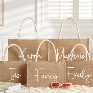 May include: A collection of burlap tote bags with white handles. Each bag is personalized with a name in white script: Karen, Marjorie, Iris, Fancy, and Emily. Pink heart-shaped sunglasses rest in front of the bags.