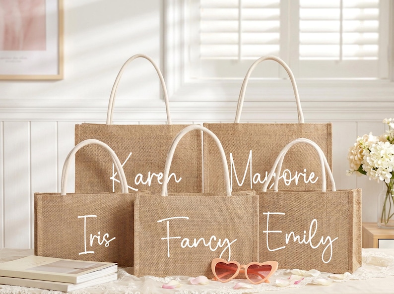 May include: A collection of burlap tote bags with white handles. Each bag is personalized with a name in white script: Karen, Marjorie, Iris, Fancy, and Emily. Pink heart-shaped sunglasses rest in front of the bags.