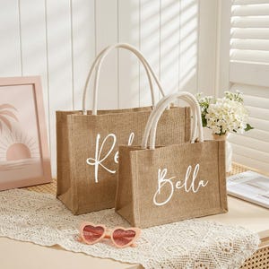 May include: Two burlap tote bags with white handles. The larger bag has the name "R" in white script, and the smaller bag has the name "Bella" in white script. The bags are sitting on a white lace tablecloth.