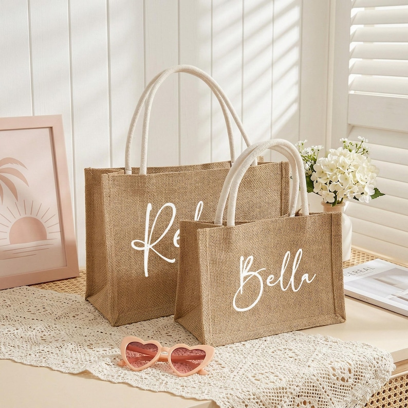 May include: Two burlap tote bags with white handles. The larger bag has the name "R" in white script, and the smaller bag has the name "Bella" in white script. The bags are sitting on a white lace tablecloth.