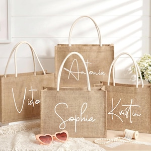 May include: Four burlap tote bags with white handles and personalized names in cursive: Vito, Antonia, Sophia, and Kristin. The bags are on a white surface with heart-shaped sunglasses and a string of pearls.