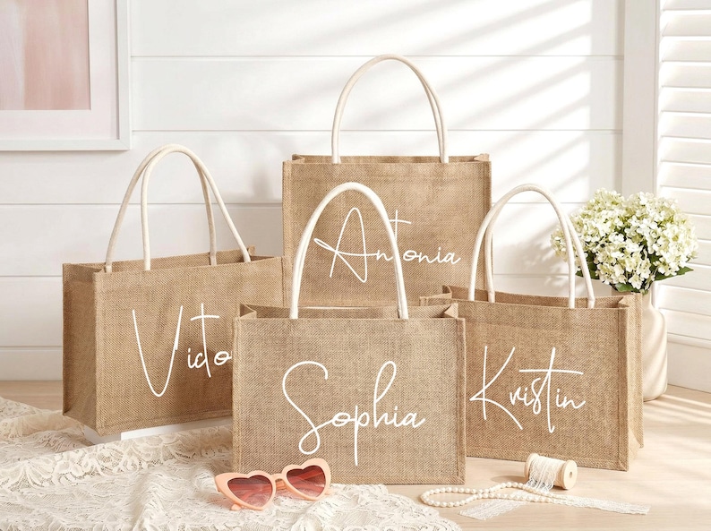 May include: Four burlap tote bags with white handles and personalized names in cursive: Vito, Antonia, Sophia, and Kristin. The bags are on a white surface with heart-shaped sunglasses and a string of pearls.