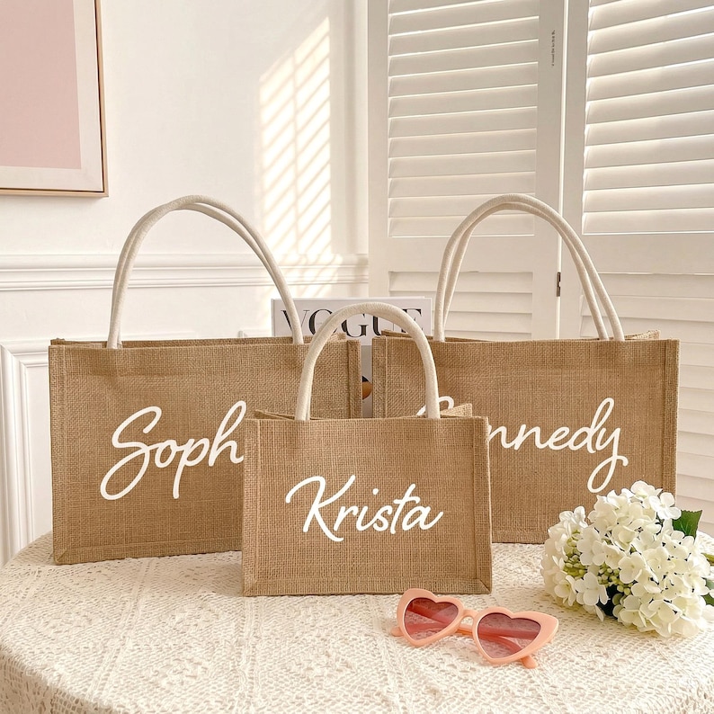 May include: Three burlap tote bags with white script names: "Soph", "Krista", and "Kennedy". The bags have white handles and are arranged on a table with a floral arrangement and pink heart-shaped sunglasses.