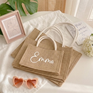 May include: A stack of three beige burlap tote bags with white handles. The top bag has the name "Emma" in white script. A pink framed picture and heart-shaped sunglasses are also in the image.