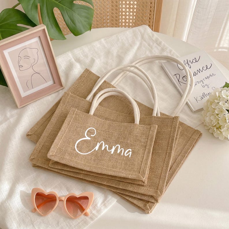 May include: A stack of three beige burlap tote bags with white handles. The top bag has the name "Emma" in white script. A pink framed picture and heart-shaped sunglasses are also in the image.