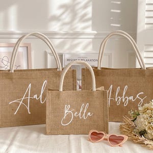 May include: Three burlap tote bags with white handles and script lettering. The bags are different sizes and have the names "Aal", "Bella", and "Abbas" written on them. Pink heart-shaped sunglasses and a bouquet of flowers are in front of the bags.