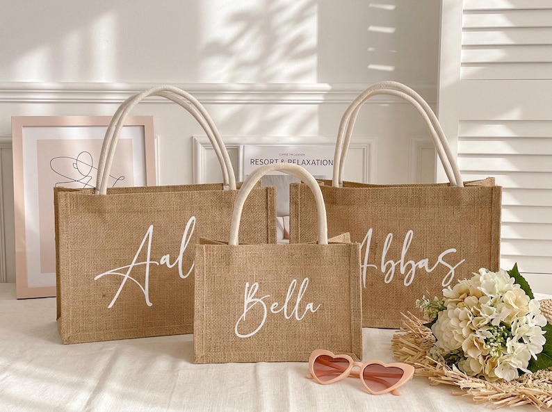 May include: Three burlap tote bags with white handles and script lettering. The bags are different sizes and have the names "Aal", "Bella", and "Abbas" written on them. Pink heart-shaped sunglasses and a bouquet of flowers are in front of the bags.