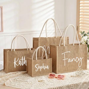 May include: Four burlap tote bags of varying sizes with white rope handles. Each bag has a white script name: Kath, Sophia, and Fancy. Pink heart-shaped sunglasses rest on the table in front of the bags.