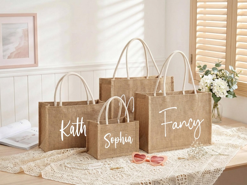 May include: Four burlap tote bags of varying sizes with white rope handles. Each bag has a white script name: Kath, Sophia, and Fancy. Pink heart-shaped sunglasses rest on the table in front of the bags.