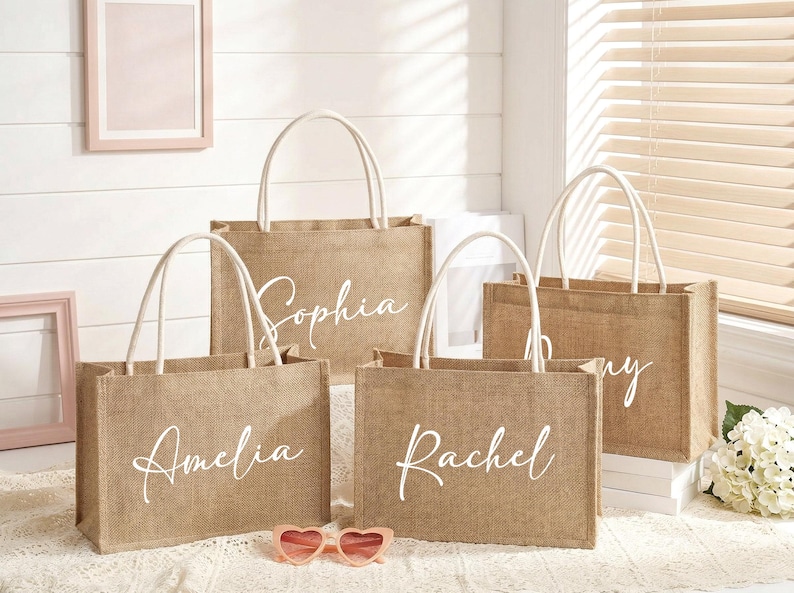 May include: Four personalized burlap tote bags with white handles. Each bag has a different name in elegant white script: Amelia, Sophia, Rachel, and Amy. Pink heart-shaped sunglasses rest in front of the bags.