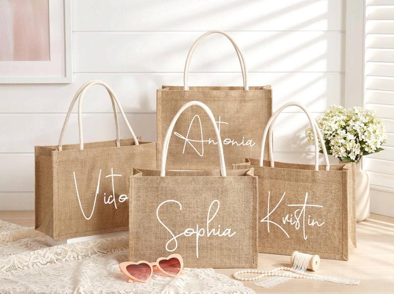 May include: Four burlap tote bags with white handles and personalized names in cursive: Vito, Antonia, Sophia, and Kristin. The bags are arranged in front of a white wall, with a vase of white flowers and a pair of pink heart-shaped sunglasses.