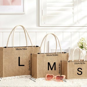 May include: Three jute tote bags in sizes L, M, and S, with measurements in inches. The bags have cream-colored handles and the letters L, M, and S printed on them. A pair of pink heart-shaped sunglasses rests in front of the bags.