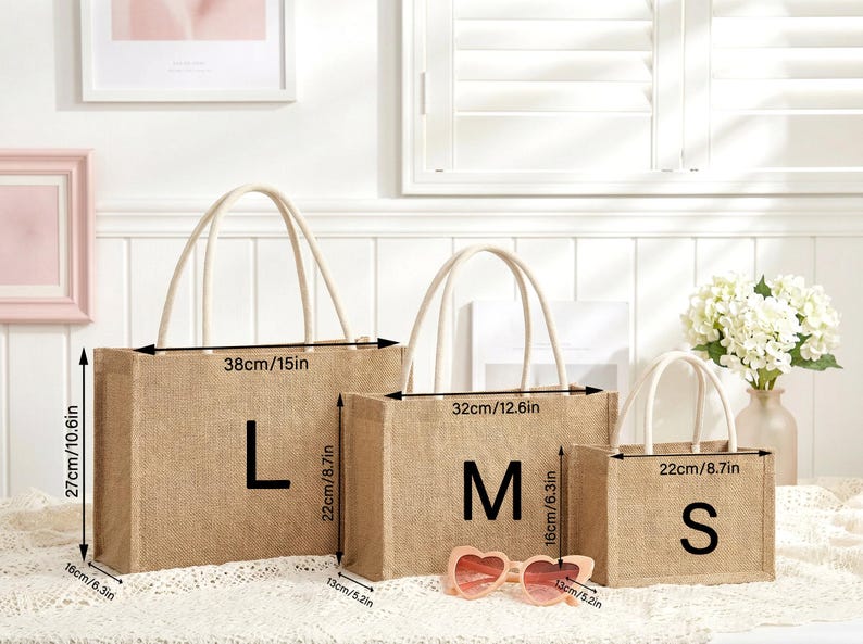 May include: Three jute tote bags in sizes L, M, and S, with measurements in inches. The bags have cream-colored handles and the letters L, M, and S printed on them. A pair of pink heart-shaped sunglasses rests in front of the bags.