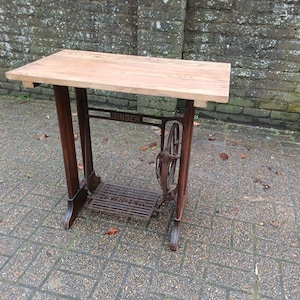 May include: A repurposed Singer sewing machine base forms a small table. It features a light wooden top and a dark brown metal base. The word "SINGER" is visible. The table stands on a brick patio, with the top measuring approximately 76 cm in length.