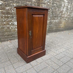 May include: A tall, rectangular wooden cabinet with a rich brown finish and a single door. The door features a recessed panel design and is equipped with a decorative, ornate metal drop handle centred on the right side of the door panel.